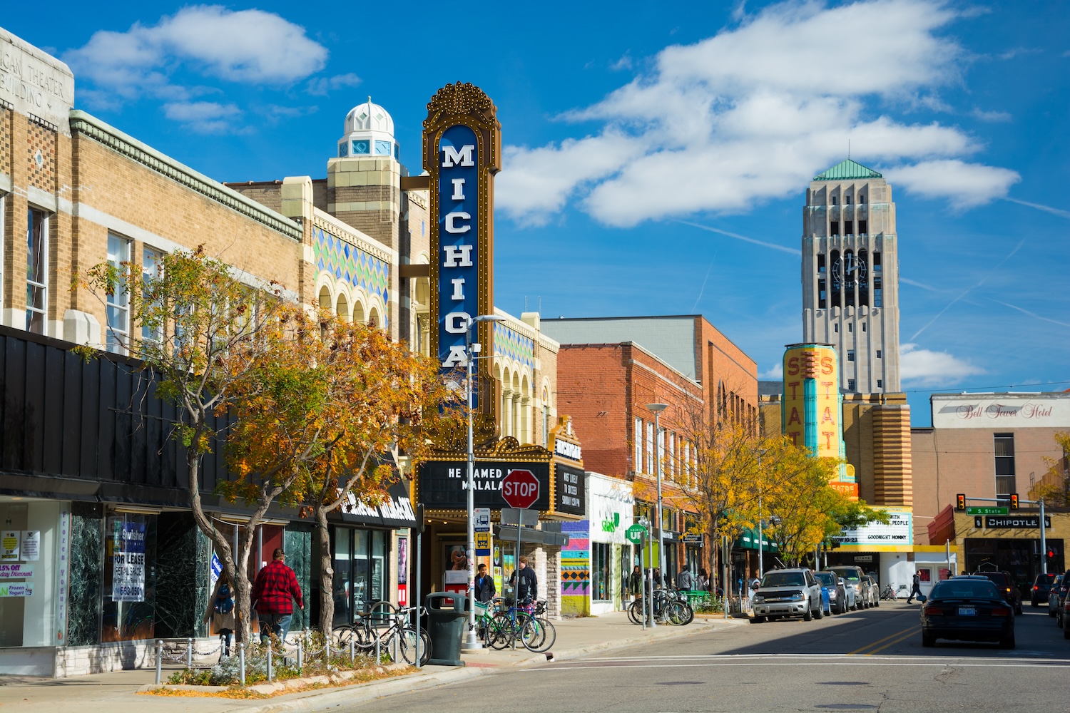 Ann Arbor, United States - October 18, 2015 - People walking in the sidewalk of Liberty Street in Downtown Ann Arbor, with storefronts and the Michigan Theater sign, State Theater sign, cars and parked bicycles in the scene, and the Burton Memorial Tower in the distance, during a day with a blue sky with clouds.
